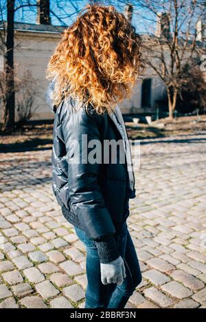 View from behind young woman with lush curly hair walking around the old town on sunny spring day. Stock Photo