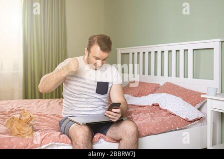 Angry young man working from home in bedroom, holding his phone in hand and a fist raised. Orange cat is standing next to him Stock Photo