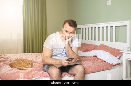 Young man working from home in bedroom, feelling bored and tired holding a phone and laptop in his arms. An orange cat is standing next to him Stock Photo