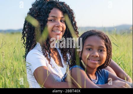 Two afro american girls smiling sitting on the grass looking at the camera. They both have long black hair. Stock Photo