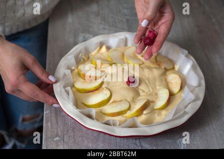 The hostess at home is preparing apple charlotte in the kitchen. puts the berry on an apple pie in a baking dish. Raw Apple Pie Dough Stock Photo