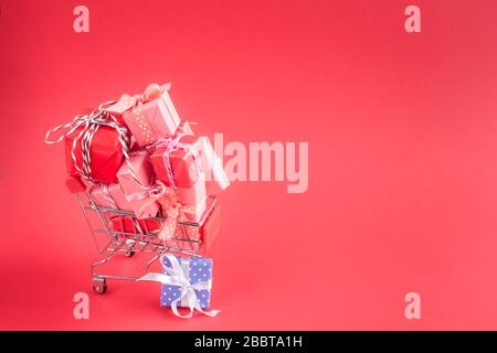 Trolley shopping cart filled with a lot of paper wrapped gift boxes on red background. Concept of online shopping. CopySpace for text. Stock Photo