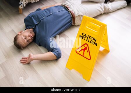 Mature Man Falling On Wet Floor In Front Of Caution Sign At Home Stock Photo