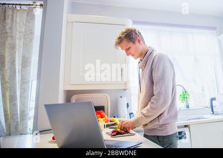 Young caucasian man working from home. Freelancer cooking vegetarian lunch on the kitchen table and working on pc laptop. Home office, remote workplac Stock Photo