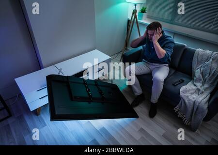 Man Sitting On Sofa In Front Fallen Television With Broken Screen Stock Photo