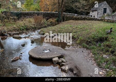 An old millstone lying in the River Dove at Viators Bridge, Milldale village, Peak District National Park, England, UK Stock Photo