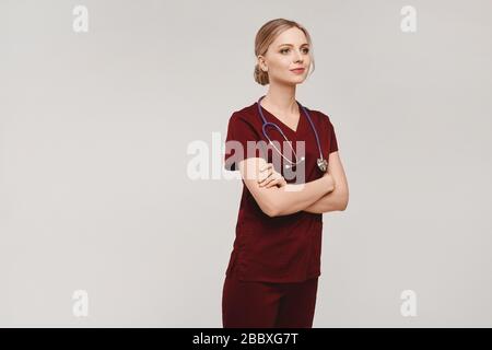 Young blonde woman in medical uniform with stethoscope posing with folded hands, isolated on a grey background. Healthcare concept Stock Photo