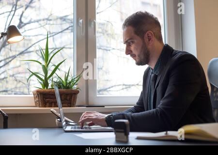 businessman working on laptop in office taking notes by window black table with notebook papers phone Stock Photo