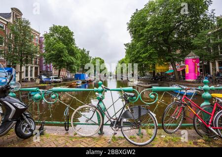 Many colored bicycles leaning agains the rail on a bridge in one of Amsterdam's canals in a rainy day. Stock Photo