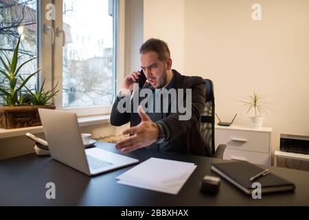 businessman working on laptop in office making phone call nervous and angry by window black table with notebook papers phone Stock Photo
