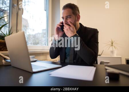 businessman working on laptop in office making phone call nervous and angry by window black table with notebook papers phone Stock Photo