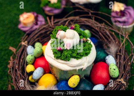 Orthodox Easter cake in a wooden nest with painted chicken and quail eggs on green grass. Easter cake with a figurine of a ponytail of a rabbit on the background of sweets, cupcakes. Stock Photo