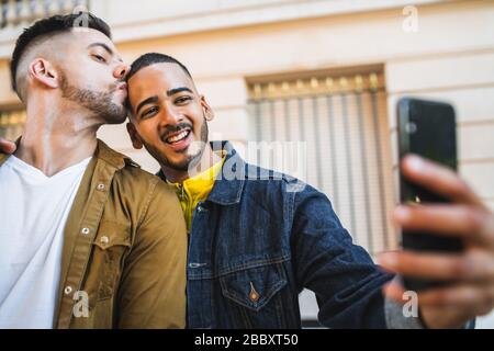 Portrait of happy gay couple spending time together and taking a selfie with mobile phone in the street. Lgbt and love concept. Stock Photo