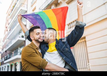 Portrait of young gay couple embracing and showing their love with rainbow flag at the street. LGBT and love concept. Stock Photo