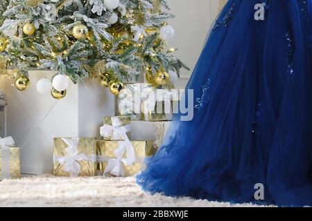 Christmas gifts under a decorated Christmas tree with white wall background. New year holiday background. Part of elegant blue dress. Stock Photo