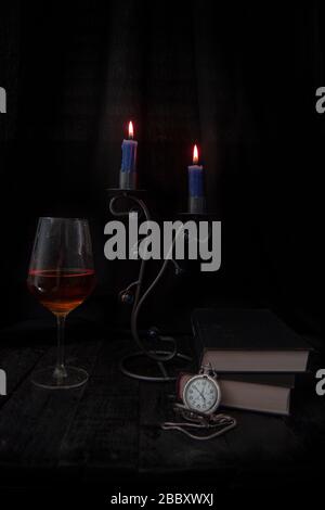 Pocket watch placed on some books, glass of red wine and candleholder with lighted candles, on black woods and black background. Stock Photo