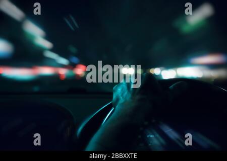 Man hands driver on steering wheel of a modern car with Car dashboard and night city background. Traffic jam on rush hour in the city. Transport Stock Photo