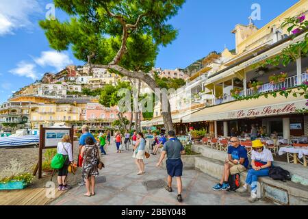 Tourists walk along the boardwalk in front of the sandy beach, cafes and shops at the coastal town of Positano Italy on the  Amalfi Coast.