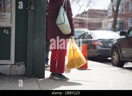 New York, NY / USA - February 28, 2020: Asian woman stands outside a corner store waiting with plastic bag in hand Stock Photo