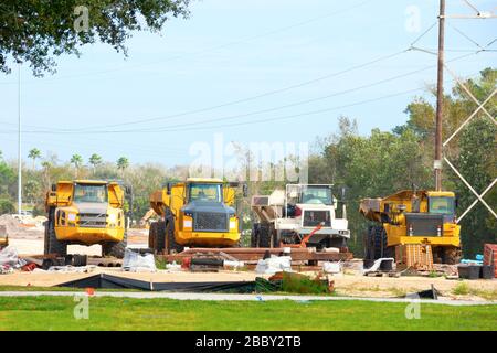 Row of working dump trucks parked in a row at construction zone site with cleared land and forrest tree line in the background. Stock Photo