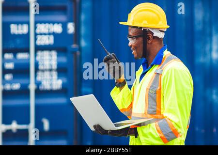Black African happy worker working in logistic communication using radio and laptop to control loading containers at port cargo to trucks for export a Stock Photo