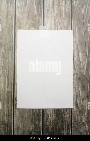 A white sheet of A4 paper hanging on a wall glued with a piece of paper tape on a wooden background. Stock Photo