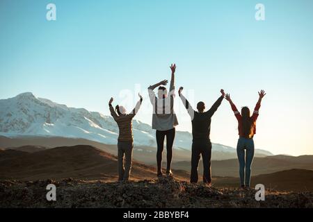 Four happy friends are standing with raised arms and looking at sunset mountains and sky. Unrecognized peoples Stock Photo