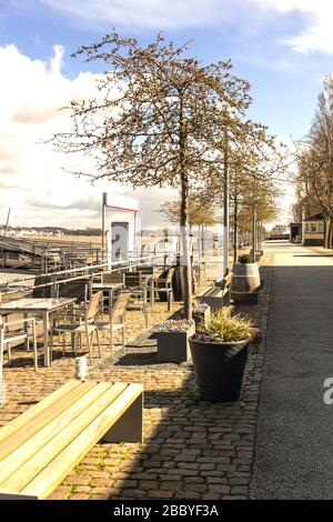 A street near the river in spring time. Sun weather. The quay with summer cafe, flower, trees, bench, tables and chairs. Stock Photo
