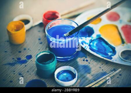 On a wooden table, there is a creative mess - in the mess are gouache paints, a palette and brushes. Stock Photo
