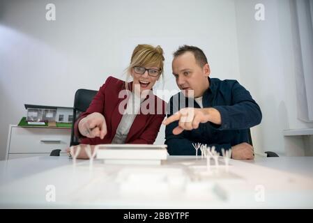 Two Architects in office sitting at desk looking over a house model. Thoughtful designers with eyeglasses thinking and discussing about project. Stock Photo