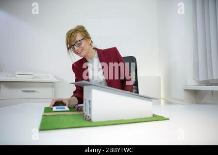Young female architect in office sitting at desk looking over a house model. Thoughtful designer with eyeglasses thinking about project. Stock Photo