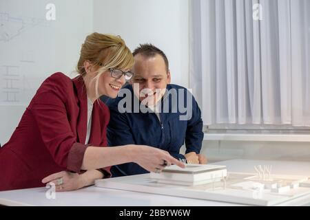 Two Architects in office sitting at desk looking over a house model. Thoughtful designers with eyeglasses thinking and discussing about project. Stock Photo