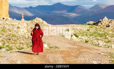 Pilgrim (woman) with red robe and hand prayer wheel walking the traditional Kora at Lake Nam Tso. Mountains of the Tibetan Plateau in the background. Stock Photo