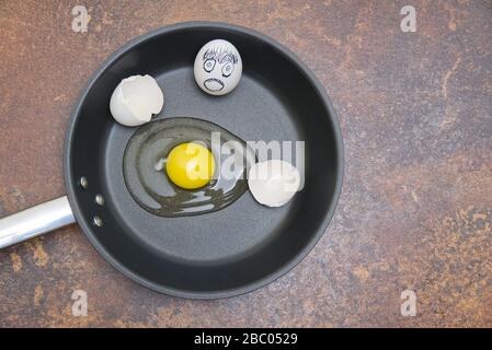 Black frying pan with raw broken egg and eggshell with emotion drawing on a rusty background Stock Photo