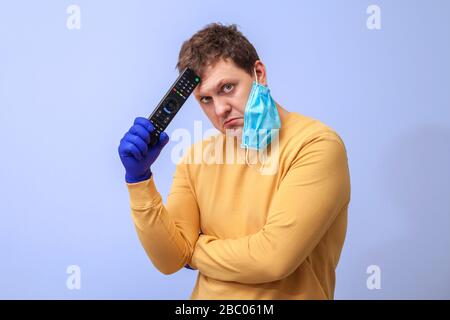 A man in protective gloves and a medical mask with a TV remote control in his hands stands with a sad expression on his face during quarantine. isolated on a blue background. concept covid-19 Stock Photo