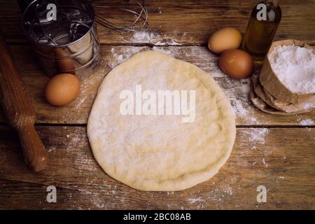 Pizza dough cooking in the home kitchen. Homemade dough for bread, pizza, pastries and rolls. Dough ingredients (flour, eggs, butter) on a wooden rust Stock Photo