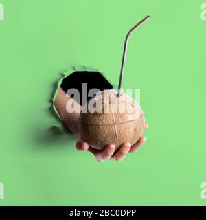 Female hand holding out one fresh coconut with metal straw inserted in it through torn hole in neo mint colored paper wall. Creative trendy concept. Z Stock Photo
