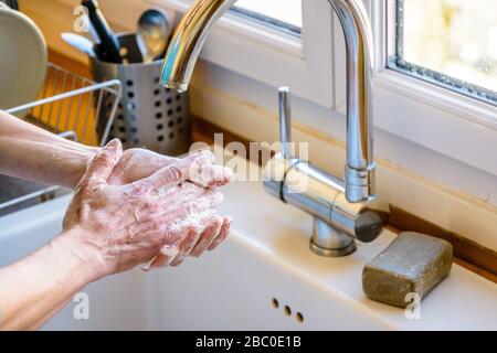 Close-up view on the hands of a woman washing her hands thoroughly with soap under the faucet of the kitchen sink.