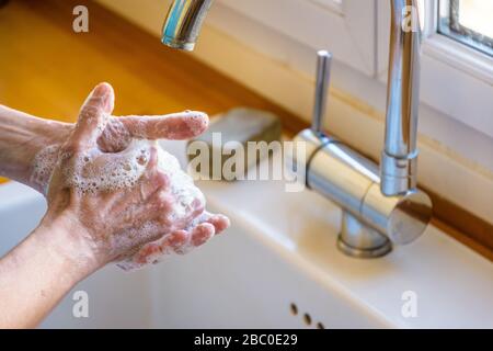 Close-up view on the hands of a woman washing her hands thoroughly with soap under the faucet of the kitchen sink.