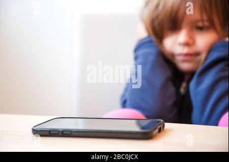 Selective focus of a smartphone on the wooden table. On the background a little girl looks the smartphone, but she don't want touch it. Childhood. Awa Stock Photo