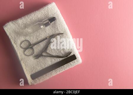 Nail saw, cuticle clippers, nail scissors and hand oil stand on a white towel that stands on a pink background. Top views Stock Photo