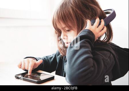 Portrait of cute little blonde girl in casual sweatshirt listening to music from smartphone with wireless headphones on the desk at home. Tech addicte Stock Photo
