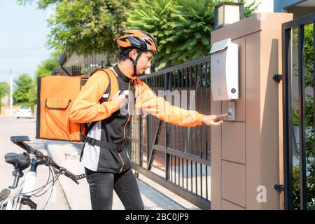 Asian delivery rider young man in orange uniform with delivery box and bicycle and ringing customers door bell in front house village with copy space. Stock Photo