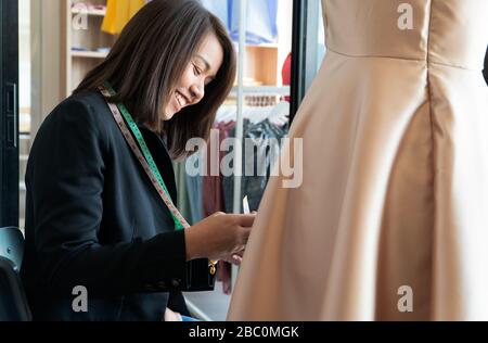 Happy young Asian woman dressmaker fashion designer is checking for completion for a suit and Dress in a showroom. Concept of success young entreprene Stock Photo