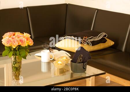 Book, glasses, roses, knitware and a mug with tea on modern glass table. Living room interior home decor concept with leather sofa. Stock Photo
