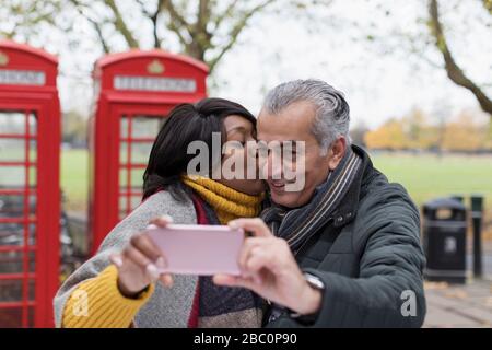 Senior couple kissing and taking selfie in park in front of red telephone booths Stock Photo