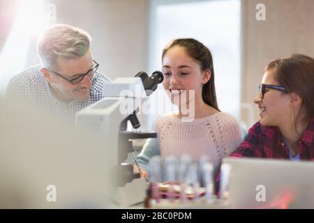 Male teacher and girl students conducting scientific experiment at microscope in laboratory classroom Stock Photo