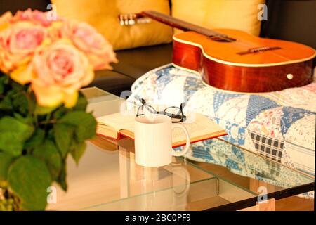 Book, glasses, roses and a mug with tea on modern glass table. An acoustic Spanish guitar is lying on an old rug on a black leather sofa. Living room Stock Photo