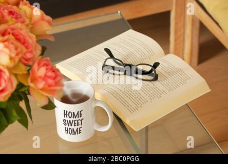 Book, glasses, roses and a mug with tea and text saying Home Sweet Home on modern glass table. Living room interior home decor concept. Selective focu Stock Photo