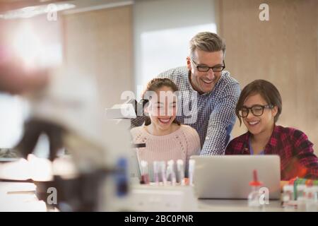 Male teacher and girl students conducting scientific experiment at microscope and laptop in laboratory classroom Stock Photo